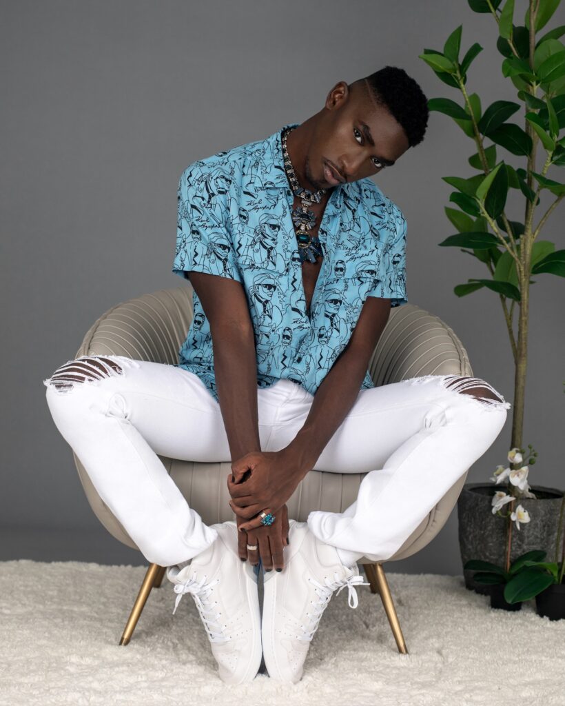 Serene portrait of a Black man in a blue button shirt and white jeans seated in a grey lounge chair — Pretty Boy Joy editorial.