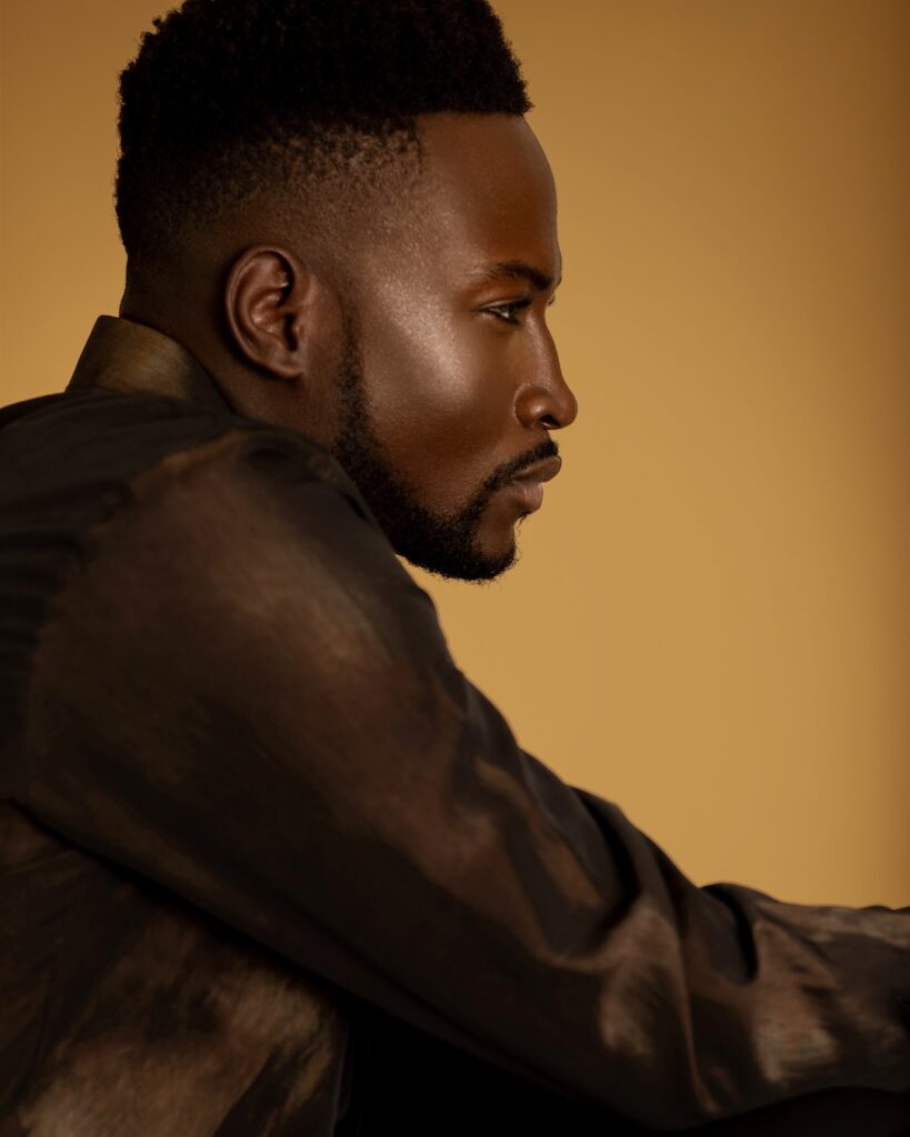 Closeup portrait of a serene Black man with short Afro hairstyle and natural makeup posing by himself inside a studio with beige background wearing a brown button up shirt.