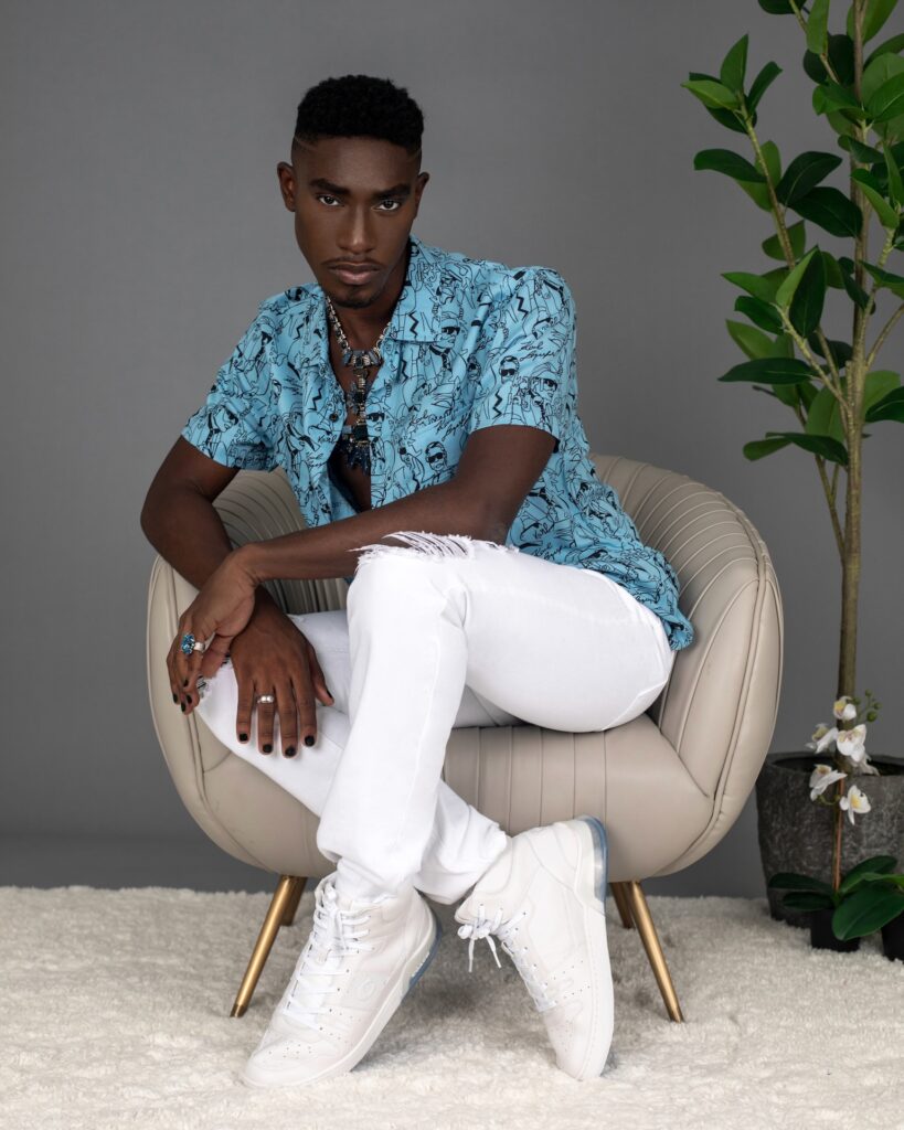 Portrait of a sensual Black man with faded Afro hair and beautiful makeup sitting by himself in a grey lounge chair indoors wearing a blue button shirt, white jeans, white sneakers and jewelry.