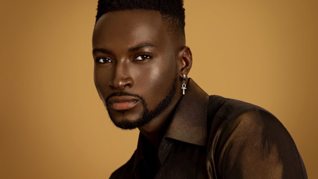 Pool Rocker Show Stopper. Closeup portrait of a confident Black man with short Afro hairstyle and natural makeup posing by himself inside a studio with beige background wearing a brown button up shirt and earring.