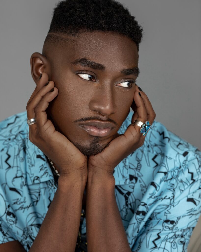 Closeup portrait of a serene Black man with faded Afro hair and beautiful makeup sitting by himself in a grey lounge chair indoors wearing a blue button shirt and jewelry.