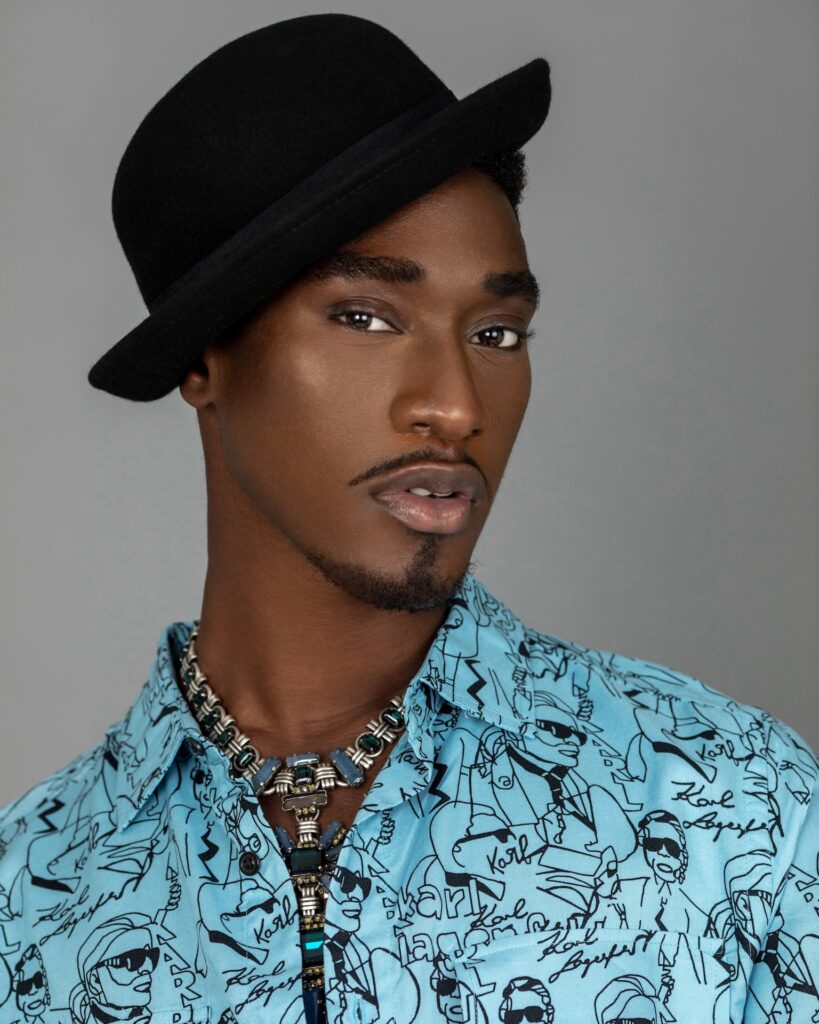 Closeup portrait of a sensual Black man with faded Afro hair and beautiful makeup sitting by himself in a grey lounge chair indoors wearing a blue button shirt with a black hat and jewelry.