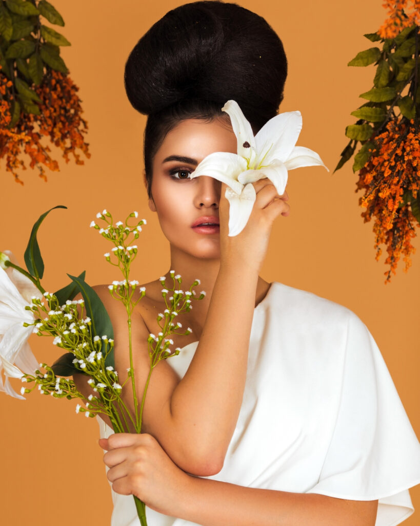Portrait of a beautiful Latin girl with long wavy hair in an up-do, beautiful makeup posing by herself surrounded by flowers inside a studio with pecan color background wearing a white evening dress.