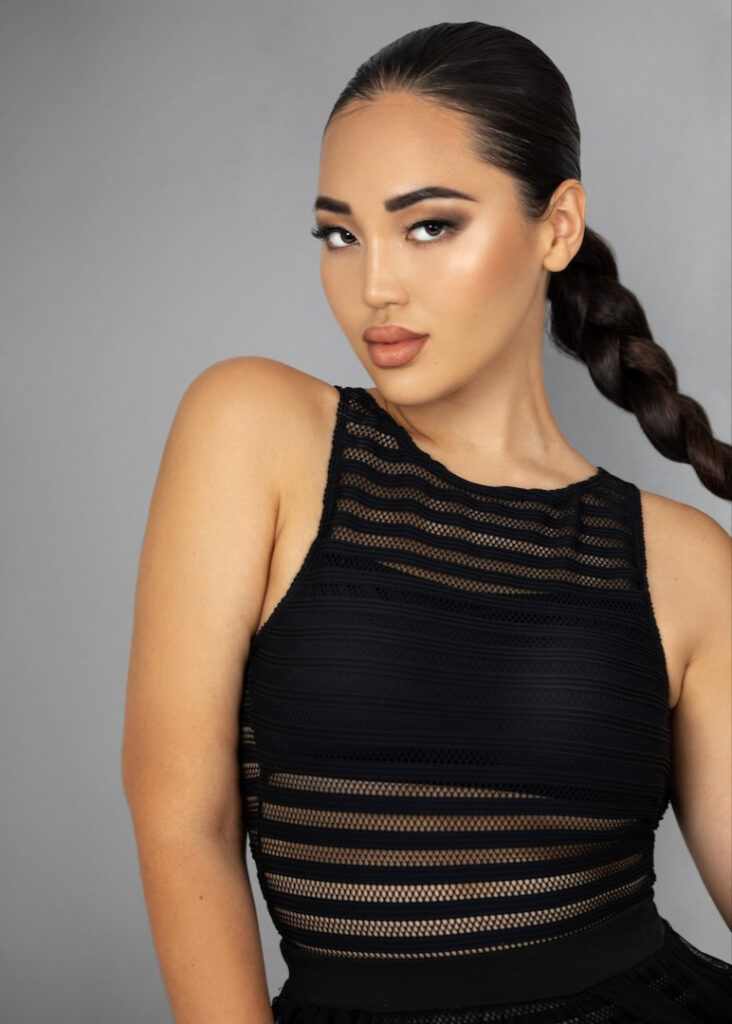 Crazy sexy confidence. Closeup portrait of a sexy young Asian woman with braided ponytail hair and beautiful makeup posing by herself inside a studio with a fading grey background wearing a black bathing suit