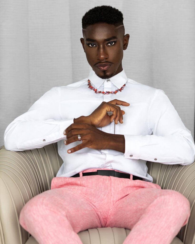 Portrait of a handsome Black man with faded Afro hair and beautiful makeup sitting in a grey leather lounge chair inside a living room wearing pink pants, a white button shirt and necklace.