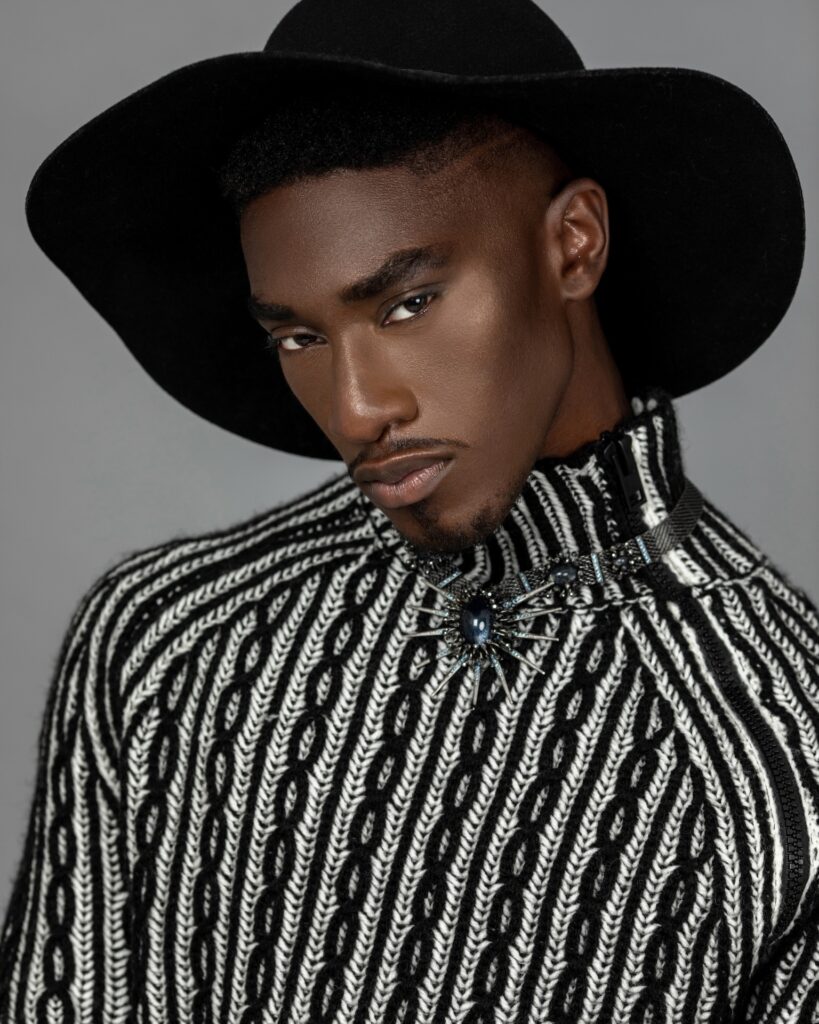 Pretty Boy Joy. Closeup portrait of a pretty Black man with faded Afro hair and beautiful makeup sitting by himself inside a studio with grey background wearing a black and white sweater, black hat and jewelry.
