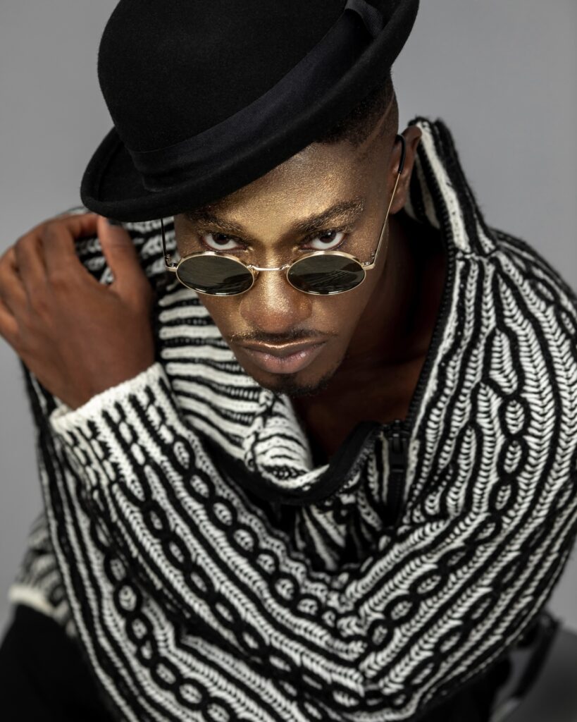 Closeup portrait of a sensual Black man with faded Afro hair and beautiful makeup sitting by himself inside a studio with grey background wearing a black and white sweater, black hat and sunglasses.
