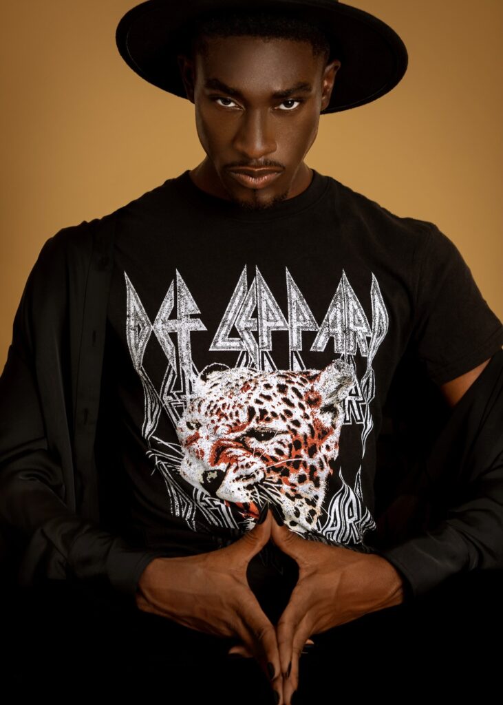 Closeup portrait of a serene Black man with fedora hat and natural makeup sitting by himself on a bar chair inside a studio with beige background wearing black pants and shirt.