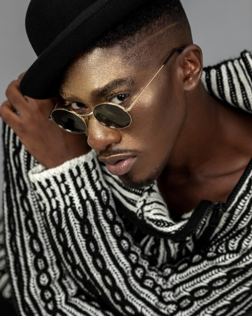 Closeup portrait of a handsome Black man with faded Afro hair and beautiful makeup sitting by himself inside a studio with grey background wearing a black and white sweater, black hat and sunglasses.