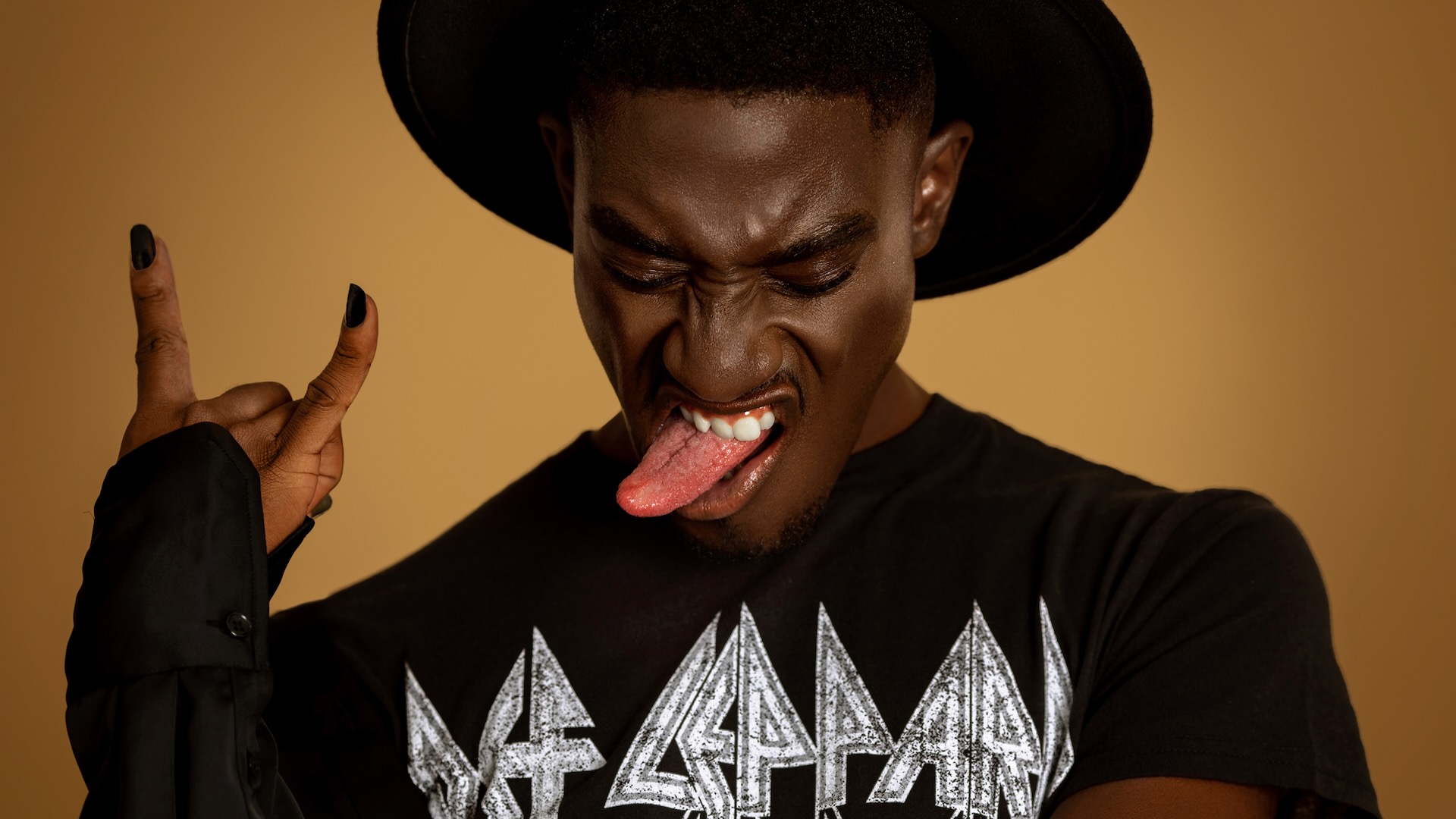 Pool Rocker Show Stopper. Closeup portrait of a serene Black man with fedora hat and natural makeup sitting by himself on a bar chair inside a studio with beige background wearing black shirt and rocking to music.