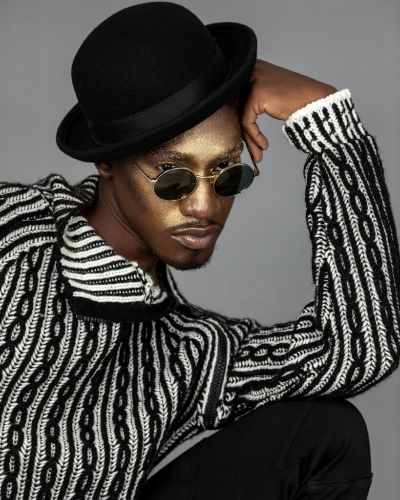 Closeup portrait of a confident Black man with faded Afro hair and beautiful makeup sitting by himself inside a studio with grey background wearing a black and white sweater, black hat and sunglasses.
