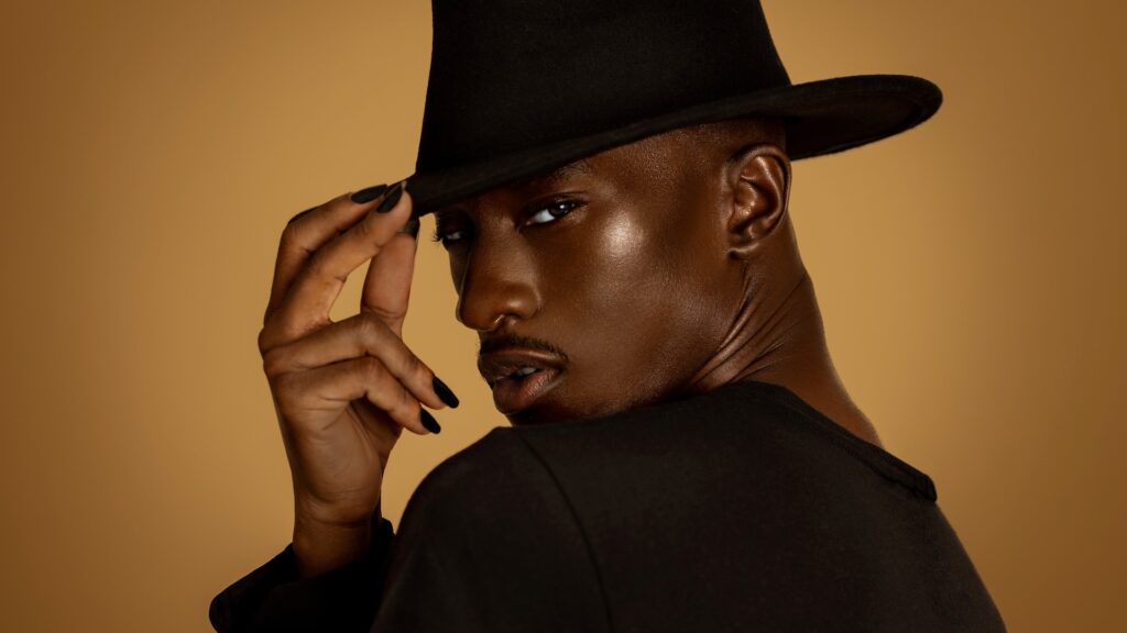 Closeup portrait of a confident Black man with natural makeup posing by himself inside a studio with beige background wearing black shirt and fedora hat tips his hat.