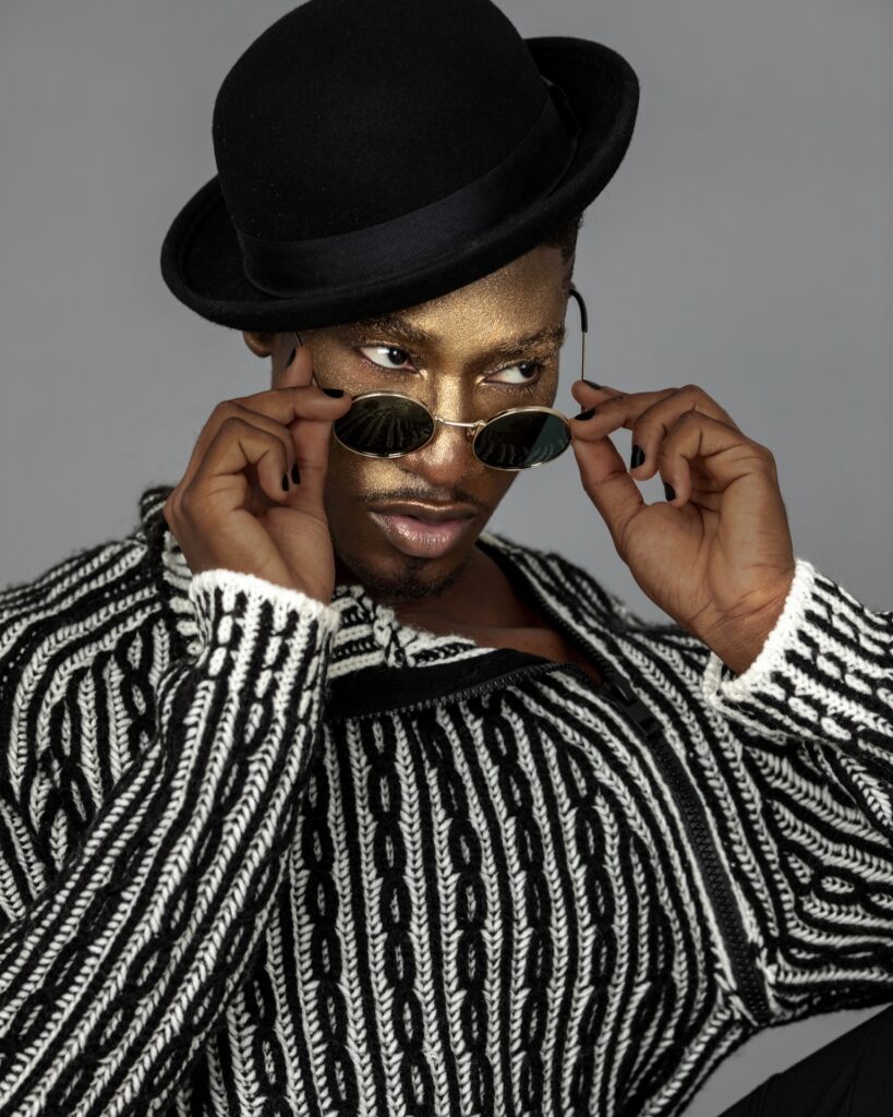 Closeup portrait of a serene Black man with faded Afro hair and beautiful makeup sitting by himself inside a studio with grey background wearing a black and white sweater, black hat and sunglasses.