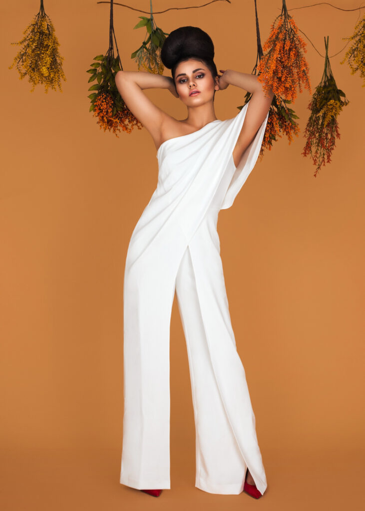 Year of self discovery. Portrait of a sensual Latin girl with long wavy hair in an up-do and beautiful makeup posing by herself surrounded by flowers inside a studio with pecan color background wearing a white evening dress.