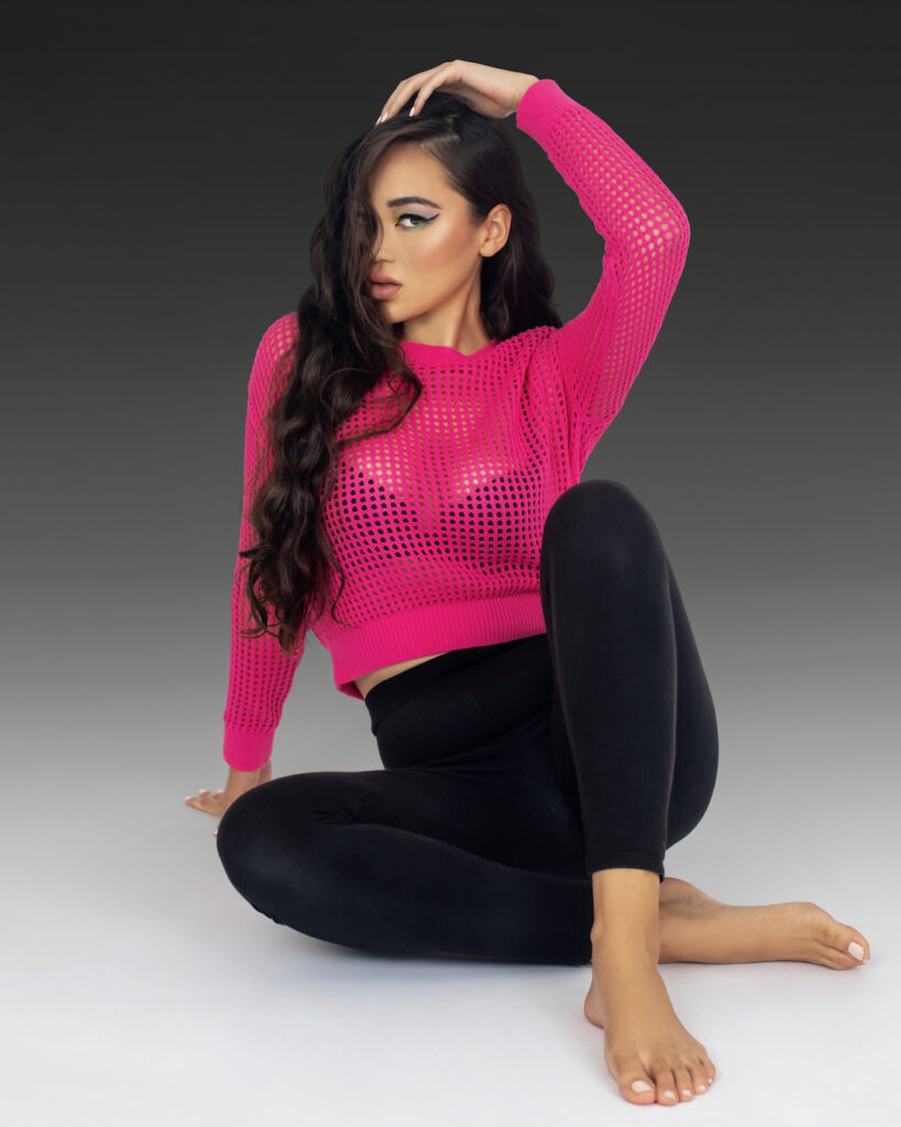 Portrait of a confident young Asian woman with long wavy hair and beautiful makeup sitting by herself on the floor inside a studio with a fading grey background wearing black pants and a pink top.