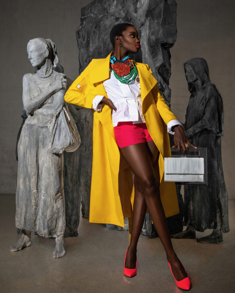 Pérez Art Museum Miami fashion editorial. An elegant young black woman with short black hair & beautiful makeup and gorgeous jewelry posing by herself indoors in front of statues is wearing a yellow coat, white blouse & red shorts.