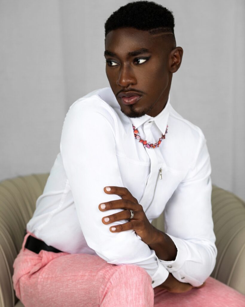 Portrait of a sensual Black man with faded Afro hair and beautiful makeup sitting in a grey leather lounge chair inside a living room wearing pink pants, a white button shirt and necklace.