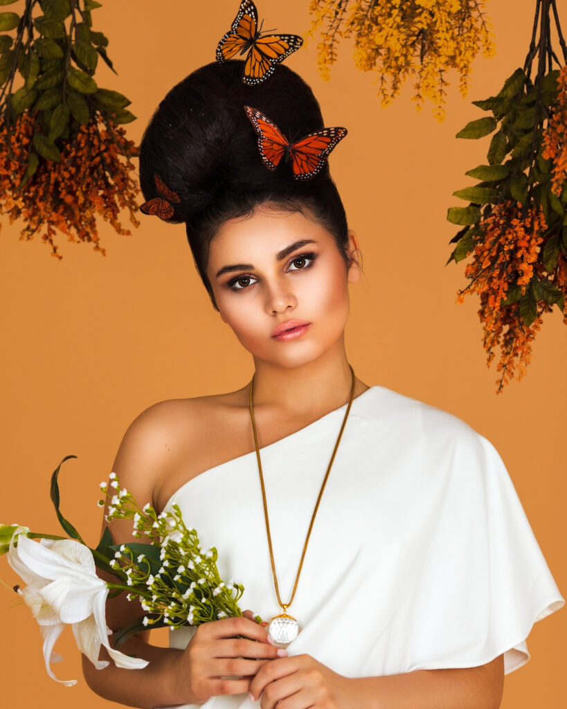 Portrait of a sensual Latin girl with long wavy hair in an up-do, beautiful makeup posing by herself surrounded by flowers inside a studio with pecan color background wearing a white evening dress.