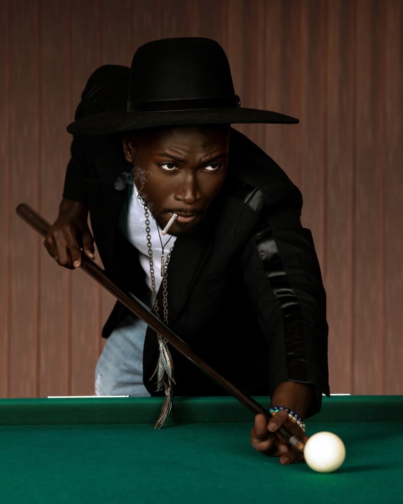 Pool Rocker Show Stopper. Closeup portrait of a focused Black man with a fedora hat posing by himself playing pool billiards wearing a black leather jacket, white T-shirt with with blue denim jeans and a long necklace.