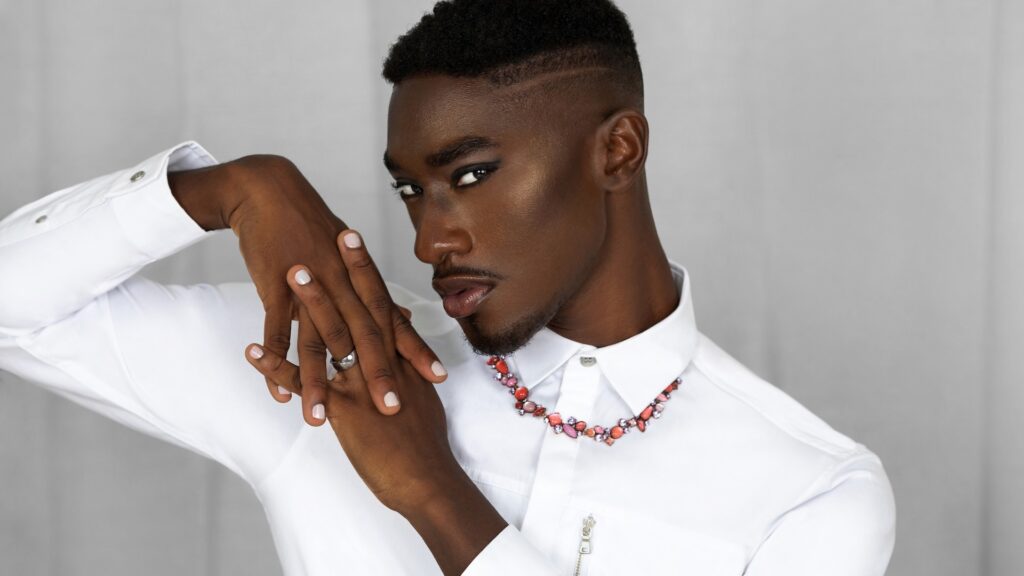 Portrait of a pretty Black man with faded Afro hair and beautiful makeup posing by himself inside a living room with a curtain background wearing a white button shirt and necklace.