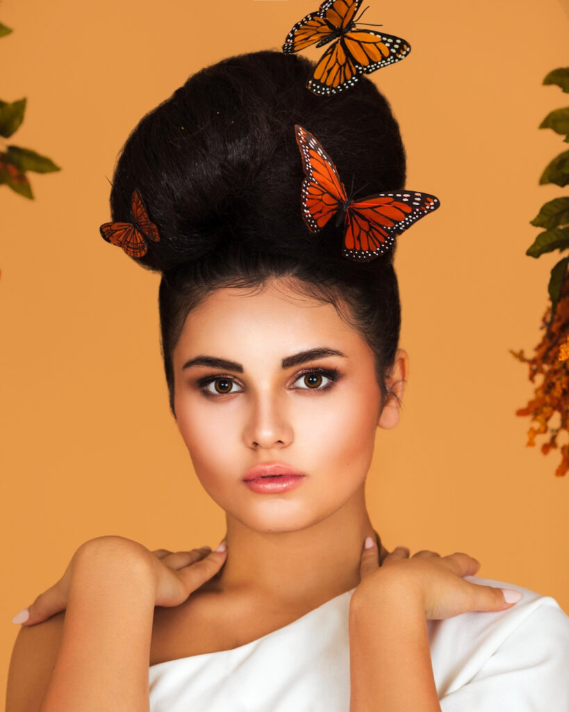 Portrait of an attractive Latin girl with long wavy hair in an up-do and beautiful makeup posing by herself surrounded by flowers and butterflies inside a studio with pecan color background.