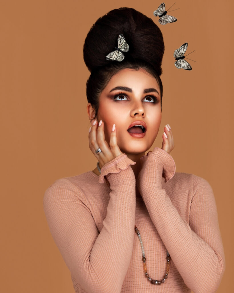 Closeup portrait of a joyful Latin girl with long wavy hair in an up-do and beautiful makeup posing by herself surrounded by butterflies inside a studio with pecan color background wearing a pink top.