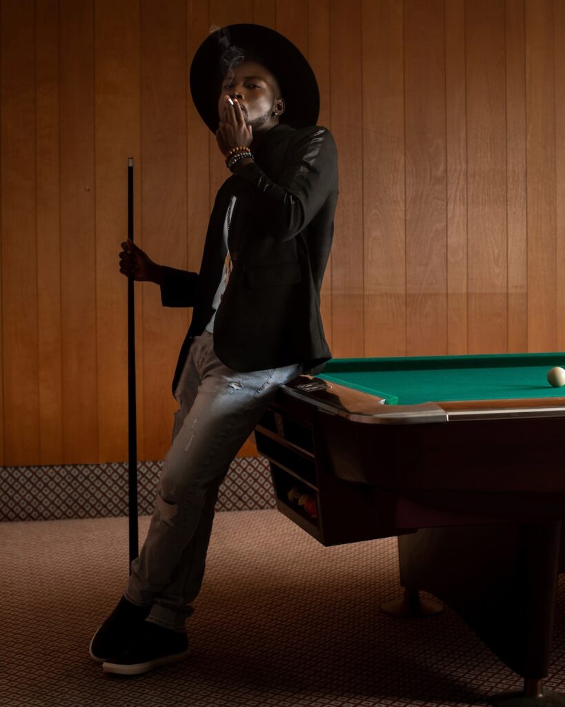 Portrait of a masculine Black man with a fedora hat posing by himself inside a pool billiards hall wearing a black leather jacket, white T-shirt with blue denim jeans and a long necklace.