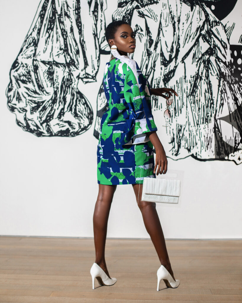 Pérez Art Museum Miami fashion editorial. An elegant young black woman with short black hair & beautiful makeup and dangling earrings posing by herself indoors is wearing a pretty dress with white high heels & a white handbag & sun glasses.