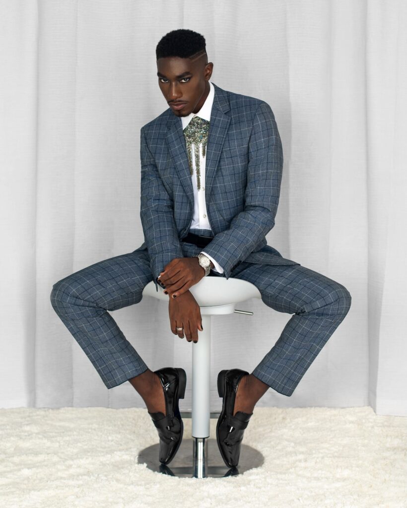 Confident Black man in a blue suit with jewelry seated on a grey bar chair — Pretty Boy Joy editorial.