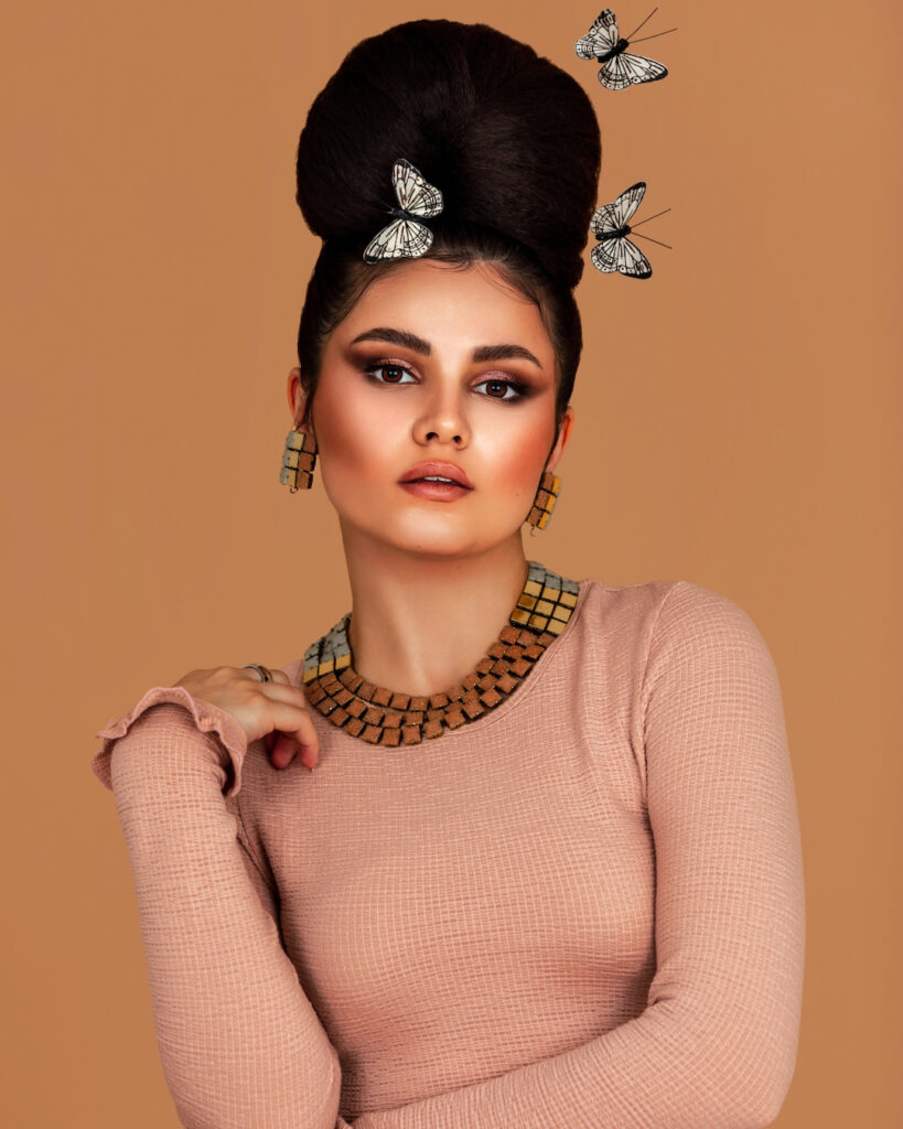 Year of self discovery. Portrait of an elegant Latin girl with long wavy hair in an up-do and beautiful makeup posing by herself surrounded by butterflies inside a studio with pecan color background wearing a pink top.