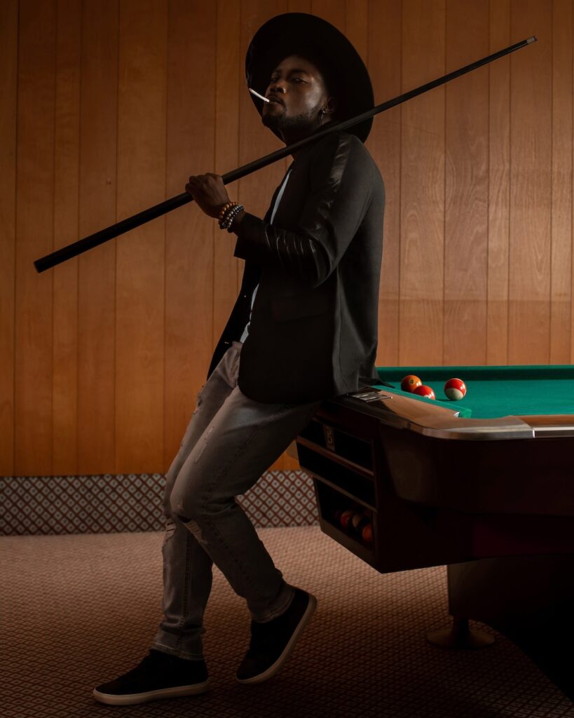 Portrait of a confident Black man with a fedora hat posing by himself inside a pool billiards hall wearing a black leather jacket, white T-shirt with with blue denim jeans and a long necklace.