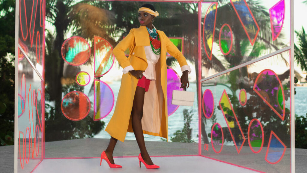 Pérez Art Museum Miami fashion editorial. An elegant young black woman with short black hair & beautiful makeup and gorgeous jewelry posing by herself outdoors inside a glass box wearing a yellow coat, white blouse & red shorts.