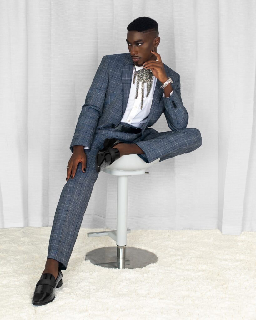 Portrait of a serene Black man with faded Afro hair and beautiful makeup sitting on a grey bar chair inside a living room with curtain background wearing blue suit, black leather shoes and jewelry.