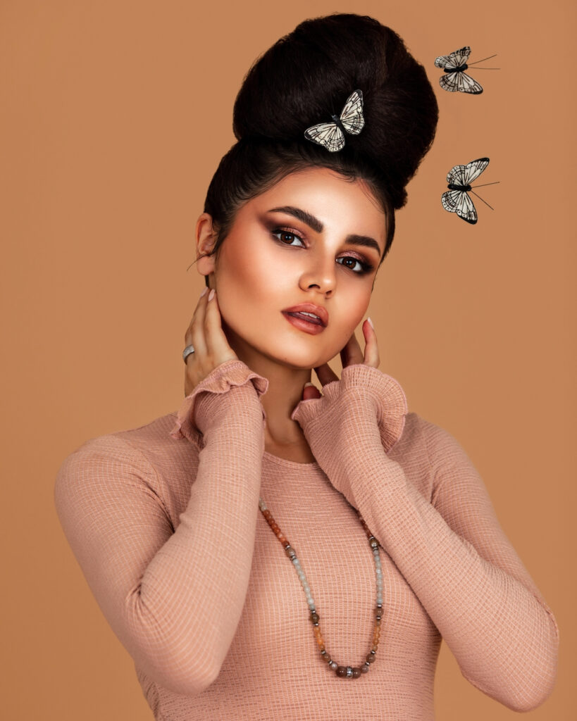 Portrait of a soulful Latin girl with long wavy hair in an up-do and beautiful makeup posing by herself surrounded by butterflies inside a studio with pecan color background wearing a pink top.