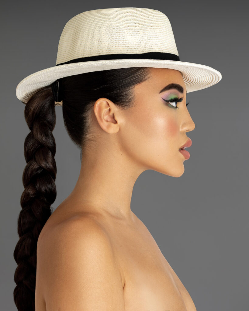 Closeup profile portrait of a serene young Asian woman with long braided ponytail hair and beautiful makeup standing by herself inside a studio with a grey background wearing a white fedora hat.