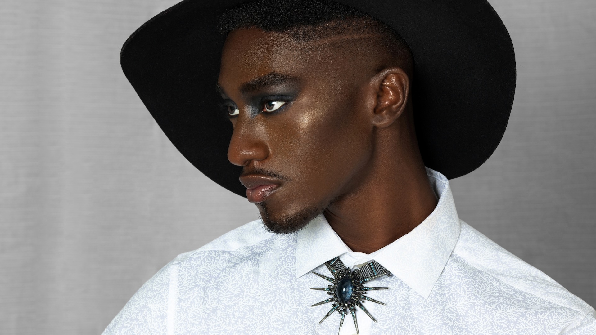 Pretty Boy Joy. Closeup portrait of a serene Black man with faded Afro hair and beautiful makeup posing by himself inside a living room with curtain background wearing a hat, white shirt and jewelry.