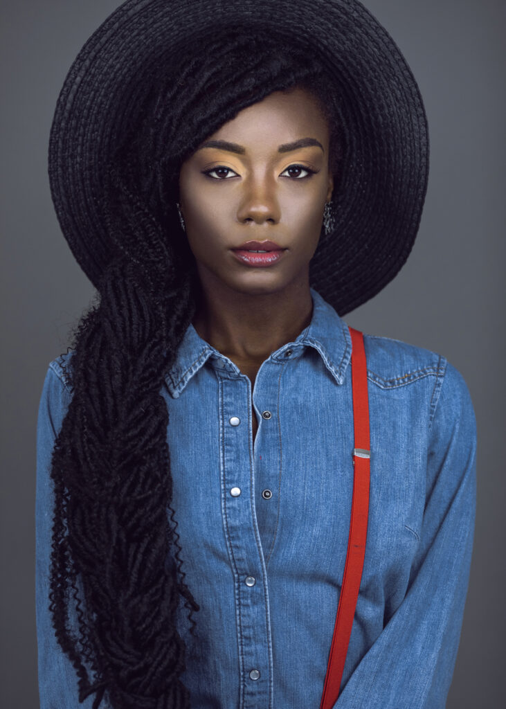 Locks of Beauty. Black female model with dreadlocks wearing denim shirt, colorful jewelry, and red suspenders in studio portrait.