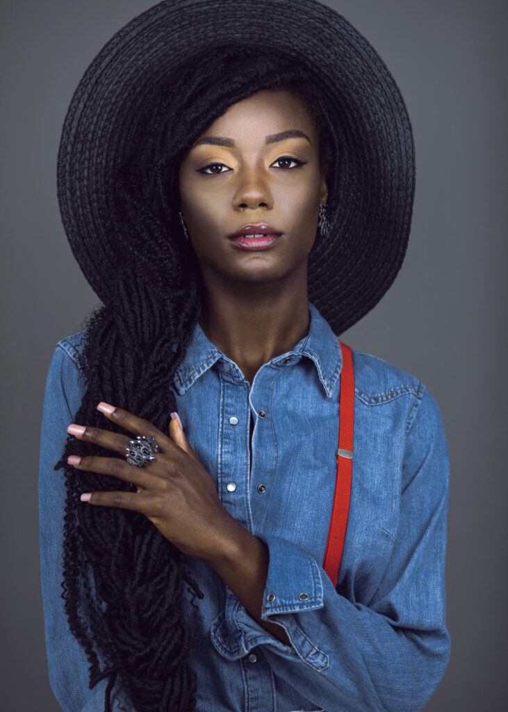 Portrait of a sensual young black female with long dreadlocks & beautiful makeup posing by herself in a studio with grey background wearing colorful jewelry, summer hat, denim shirt & red suspenders.