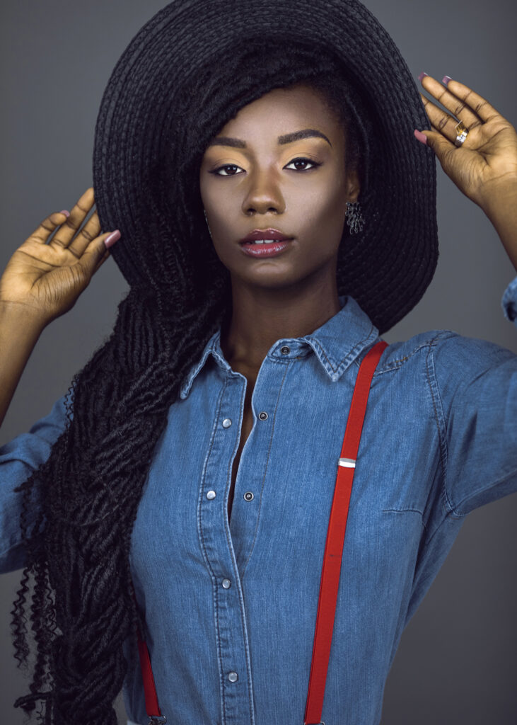 Portrait of a sexy young black female with long dreadlocks & beautiful makeup posing by herself in a studio with grey background wearing colorful jewelry, summer hat, denim shirt & red suspenders.