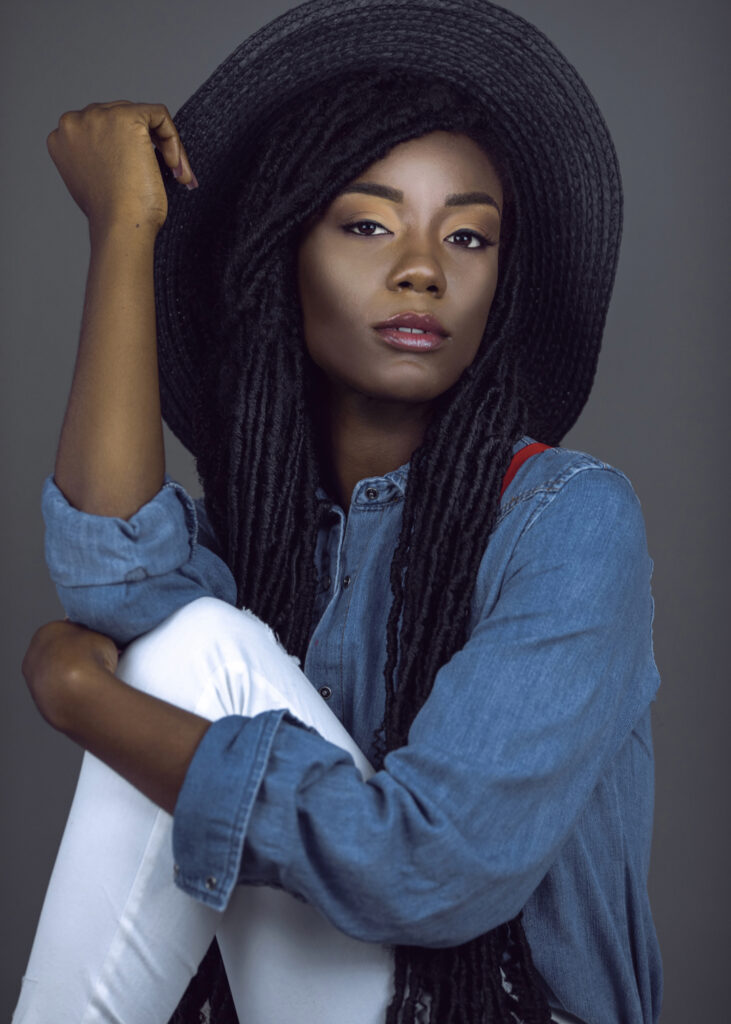 Portrait of a beautiful young black female with long dreadlocks & beautiful makeup posing by herself in a studio with grey background wearing colorful jewelry, summer hat, denim shirt & red suspenders.