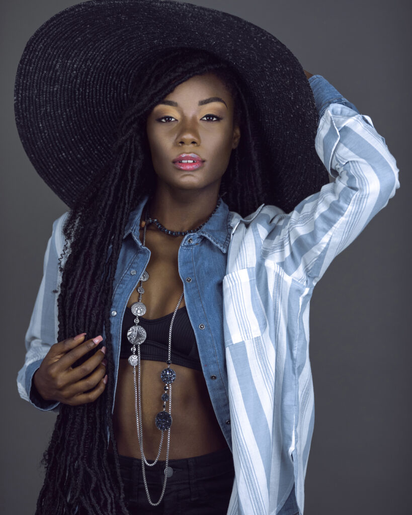 Portrait of a beautiful young black female with long dreadlocks, beautiful makeup, moist lips posing by herself in a studio with grey background wearing a summer hat & outfit with jewelry.