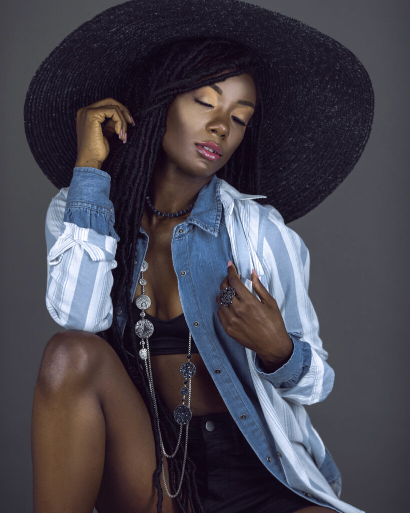 Portrait of a serene young black female with long dreadlocks, beautiful makeup, moist lips posing by herself in a studio with grey background wearing a summer hat & outfit with jewelry