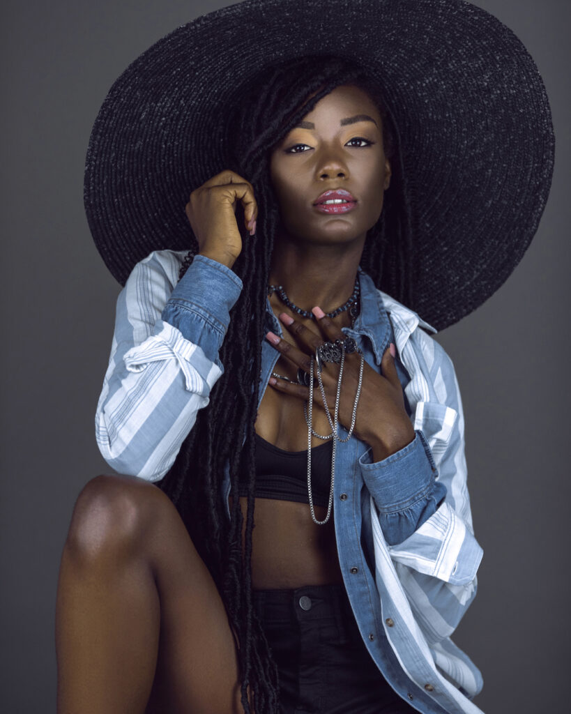 Portrait of a sensual young black female with long dreadlocks, beautiful makeup, moist lips posing by herself in a studio with grey background wearing a summer hat & outfit with jewelry.