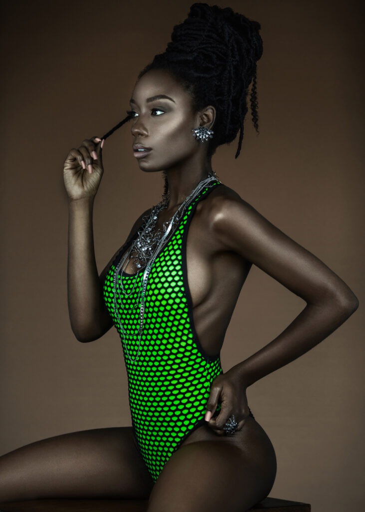 Portrait of a serene young black female with long dreadlocks, beautiful makeup and moist lips posing by herself in a studio with brown background wearing jewelry & a green bathing suit.