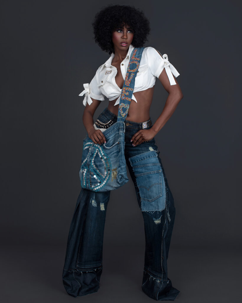 Portrait of a sexy Black woman with curly black hair and beautiful makeup standing by herself inside a studio with grey background wearing denim jeans and bag with white shirt and black high heels.