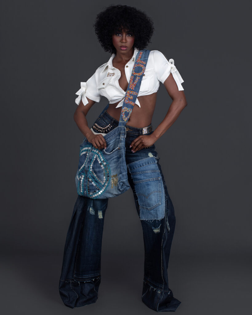 Portrait of a confident Black woman with curly black hair and beautiful makeup standing by herself inside a studio with grey background wearing denim jeans and bag with white shirt and black high heels.