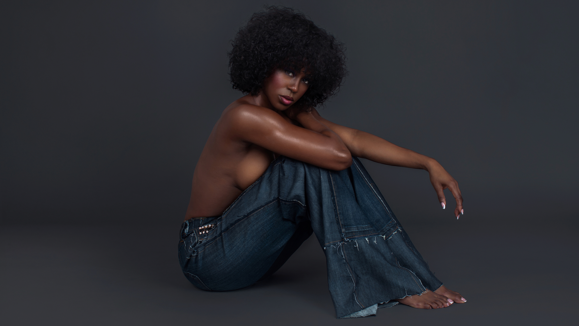 The Exhibitionist fashion editorial. Portrait of a sensual Black woman with curly black hair and beautiful makeup sitting by herself inside a studio with grey background wearing denim jeans.