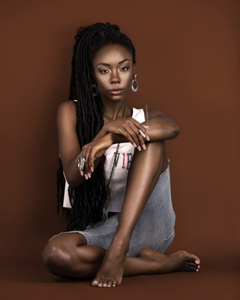 Portrait of a sexy black female with long dreadlocks, beautiful makeup, moist lips sitting on the floor by herself in a studio with brown background wearing a summer outfit while holding a joint.