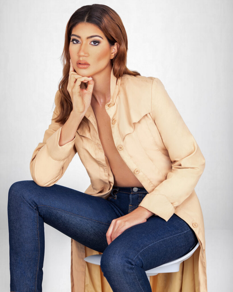 Portrait of a sensual young Latina with long wavy hair and beautiful makeup sitting on a bar chair inside a studio wearing a beige coat and blue denim jeans.