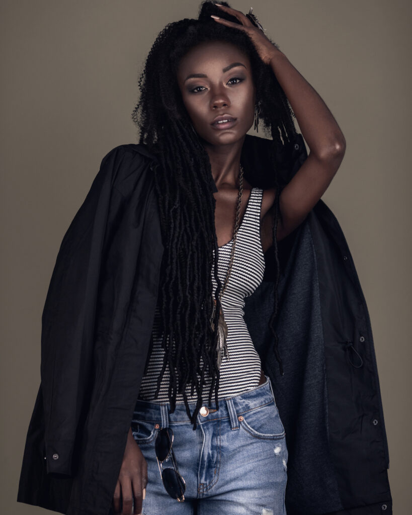 Portrait of a young black female with long dreadlocks, beautiful makeup and moist lips posing by herself in a studio with brown background wearing jewelry, denim shorts, a body top & jacket.